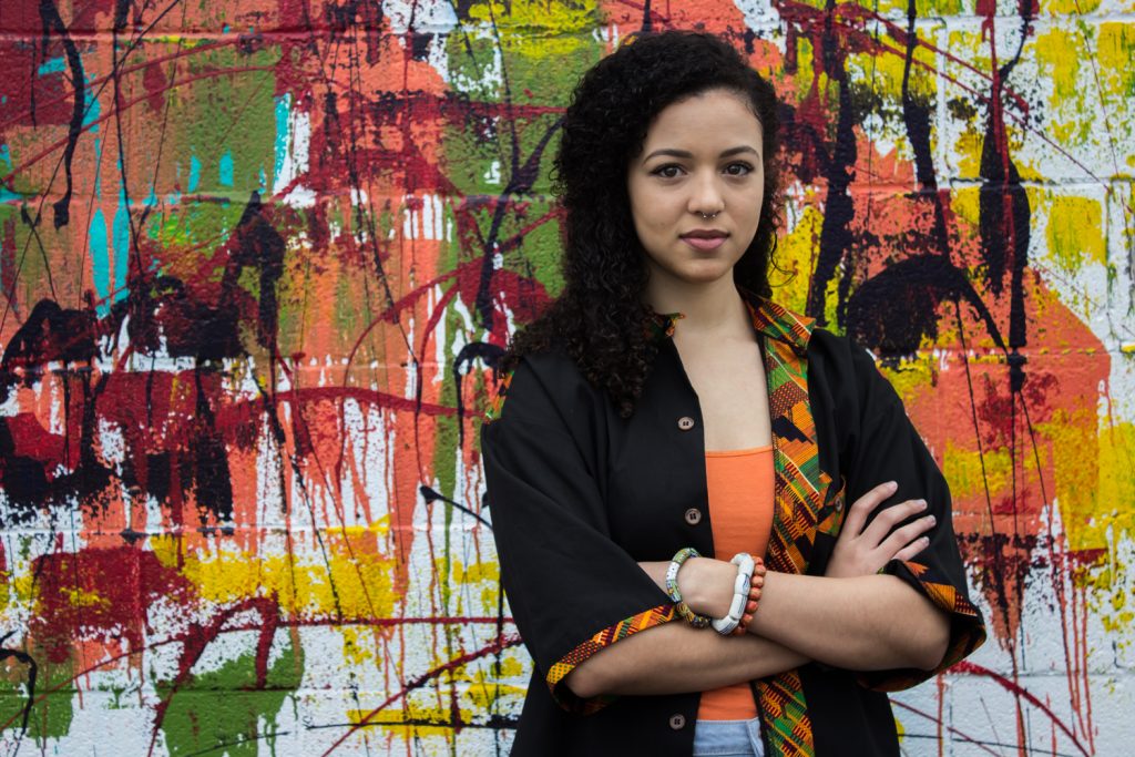 A woman is crossing her arms in front of a wall with spray paint and graffiti.