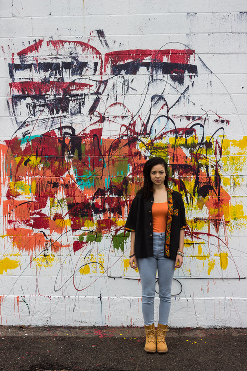 A woman stands in front of a wall with graffiti and spray paint on it.
